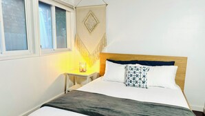 Un lit avec une tête de lit en bois, une table de chevet avec une lampe, une décoration murale et une fenêtre avec des stores.