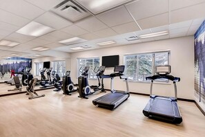 Uma academia com diversos equipamentos de exercício, grandes janelas e um espelho refletindo o espaço.