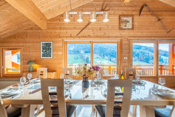A wooden interior with a dining table set for four, large windows with a view of a forested landscape, and a rustic ceiling with exposed beams. - 4th floor, balcony, parking, ski locker, 132m², Le Grand Bornand (Le Grand-Bornand)