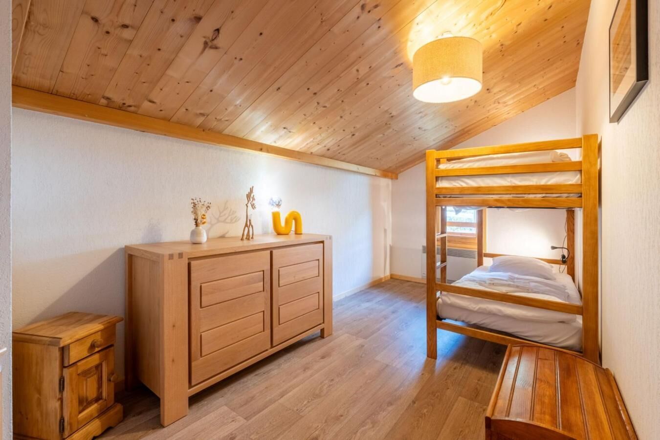 Une chambre avec un lit superposé, une commode en bois et un luminaire au plafond.