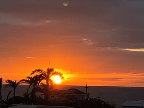 Ein Sonnenuntergang über einer Küstenlandschaft mit Palmen und einem Haus im Vordergrund.