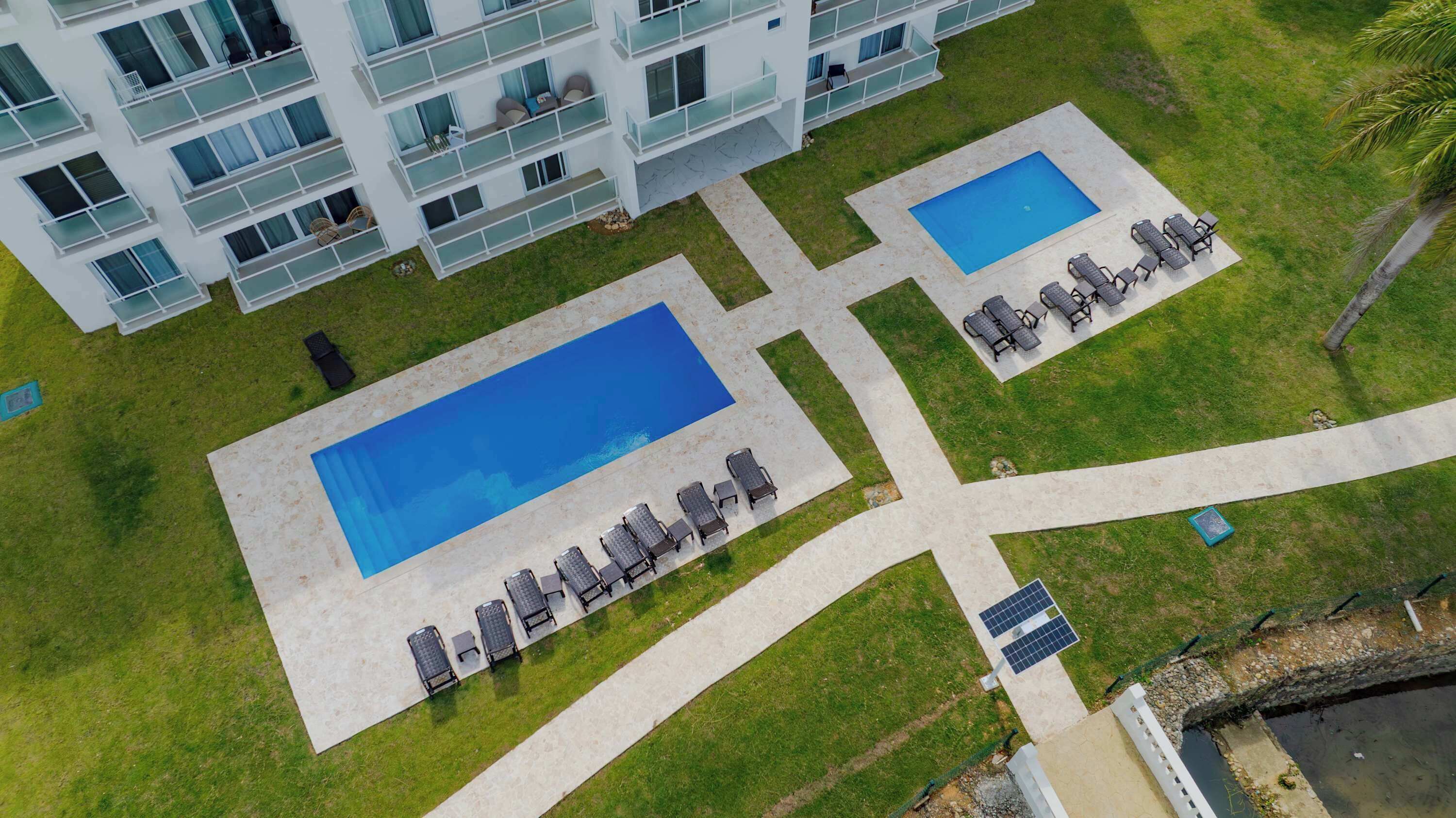 Vue aérienne d’un hôtel doté de deux piscines, de chaises longues et d’un bâtiment moderne avec des balcons.