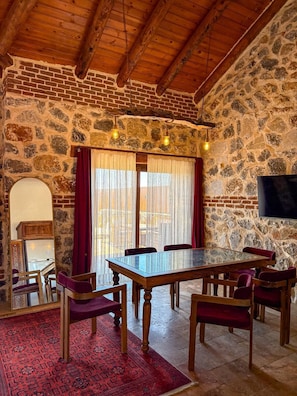 Una habitación con pared de piedra, vigas de madera en el techo, una gran mesa con sillas, un televisor de pantalla plana y un tapete rojo con estampado.