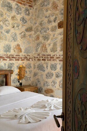 Una habitación con pared de piedra, cabecera de madera, mesita de noche de madera y cama con ropa blanca.