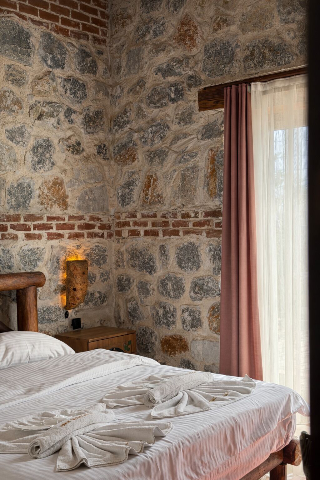 Un dormitorio con pared de piedra, cabecero de madera, mesita de noche con lámpara y ventana con cortinas.