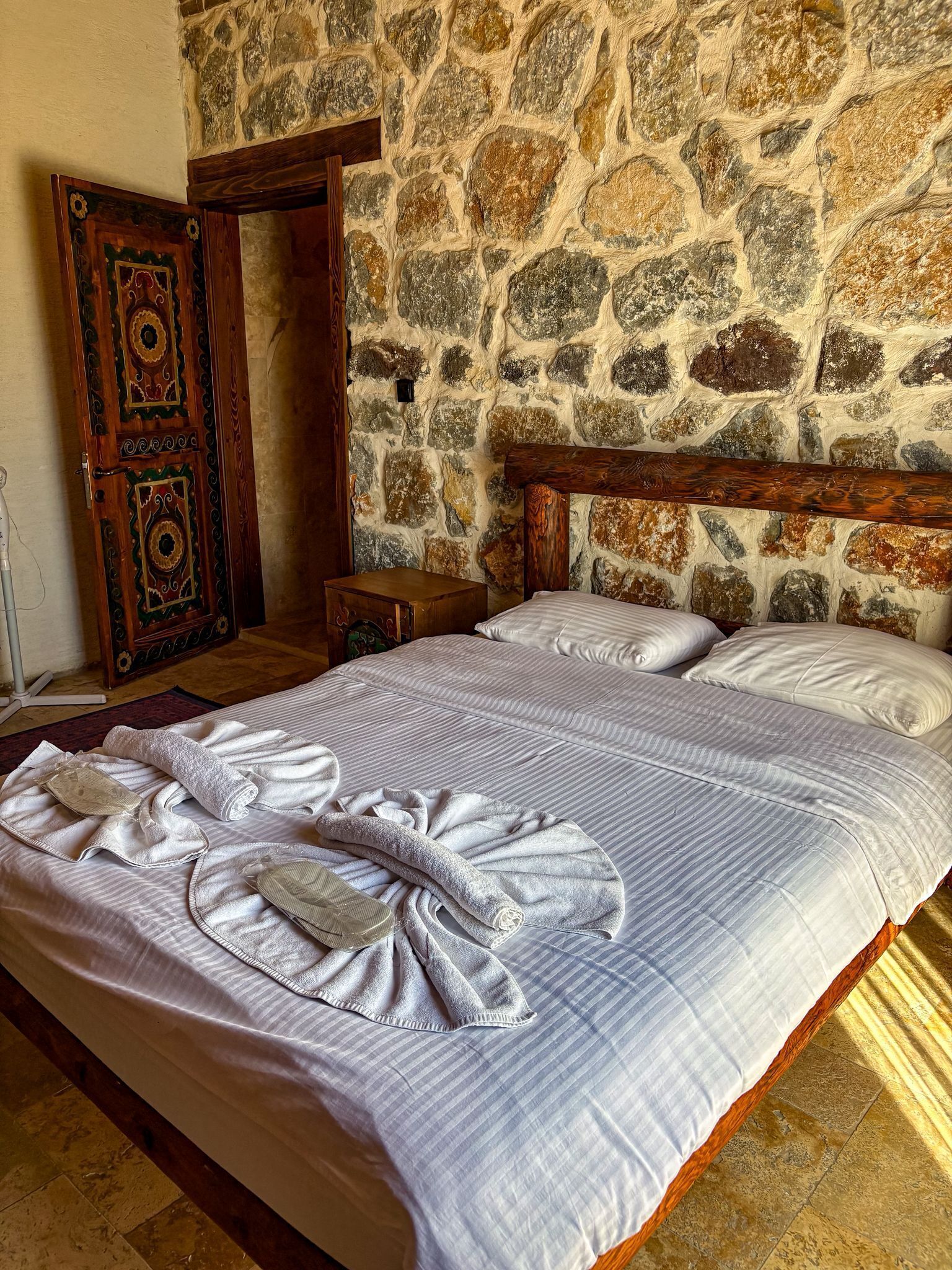 Une chambre avec un mur en pierre, une porte en bois ornée de motifs complexes, un lit avec des serviettes soigneusement disposées et une petite table de chevet en bois.