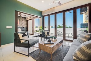 A modern living room with a sofa, armchair, coffee table, and large windows offering a view of the outdoors. - Zona Romantica Condo | Rooftop Pool | 1 Min Beach (Puerto Vallarta)