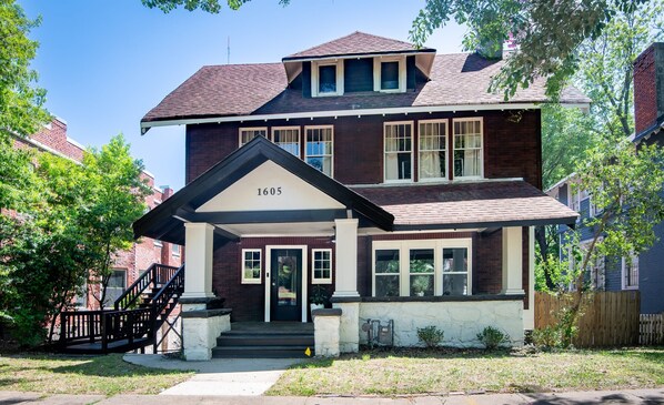A two-story brick house with a front porch, a number '1605' above the door, and a small garden area.