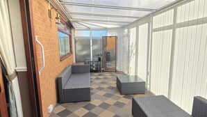 Une véranda avec un sol à carreaux, une porte d’entrée en verre et un coin salon avec des coussins.