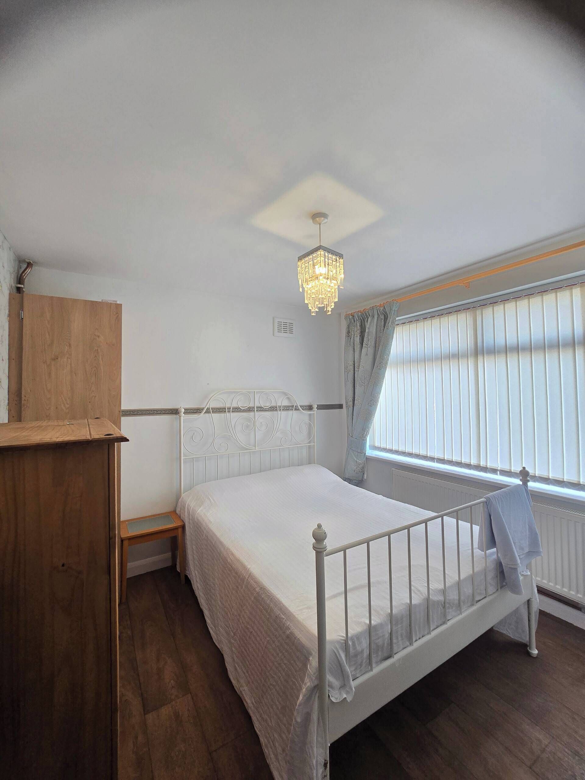 Een slaapkamer met een houten garderobe, een wit bed met metalen frame, een kroonluchter en een raam met gordijnen.