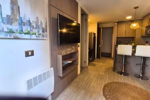 A modern living room with a flat-screen TV, built-in shelves, a wooden floor, and a cityscape painting on the wall. - Vista Ñuñoa | 1 Minute Subway | Gym | Cowork | Parking (Ñuñoa)