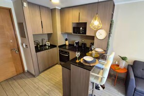 A compact kitchen with wooden cabinets, a black countertop, and a dining area with a white chair and a small orange table. - Vista Ñuñoa | 1 Minute Subway | Gym | Cowork | Parking (Ñuñoa)