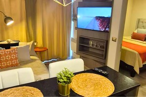 A hotel room with a sofa, a dining table with a remote control, a television, and a bed with a red blanket. - Vista Ñuñoa | 1 Minute Subway | Gym | Cowork | Parking (Ñuñoa)