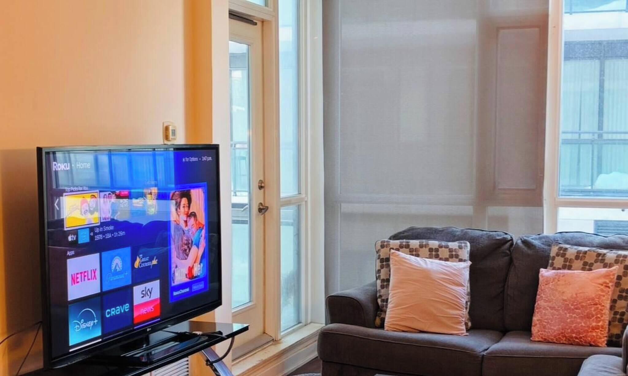 Ein Fernseher zeigt verschiedene Streaming-Dienste, ein Sofa mit gemusterten Kissen und ein Fenster mit Blick.