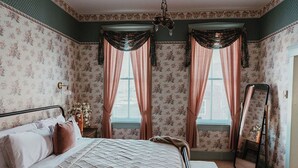 Un dormitorio con cama, cortinas, un espejo y papel tapiz con motivos florales.