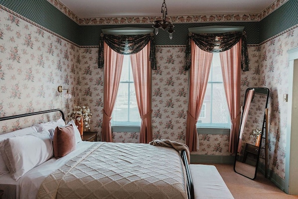 A bedroom with a bed, curtains, a mirror, and floral wallpaper.