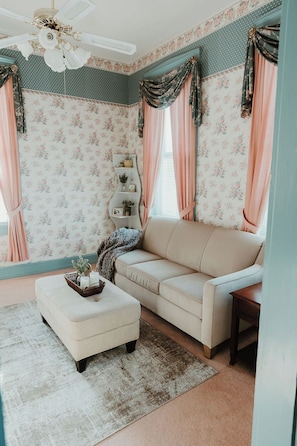 A living room with floral wallpaper, a beige sofa, an ottoman, a ceiling fan, and a window with curtains.