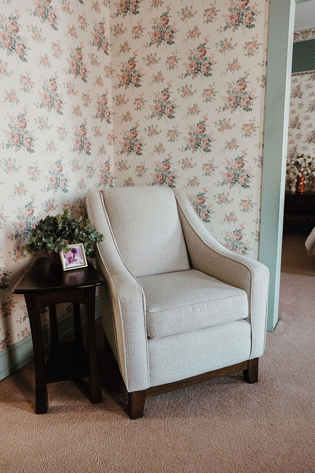 Un fauteuil de couleur claire, doté d’un coussin, est placé à côté d’une petite table sur laquelle se trouvent une plante et un cadre photo. La pièce est décorée de papier peint à motifs floraux et le sol est recouvert d’une moquette.
