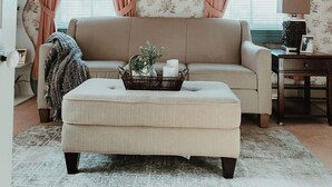 Una sala de estar con un sofá color beige, un reposapiés, una mesita auxiliar y una lámpara.