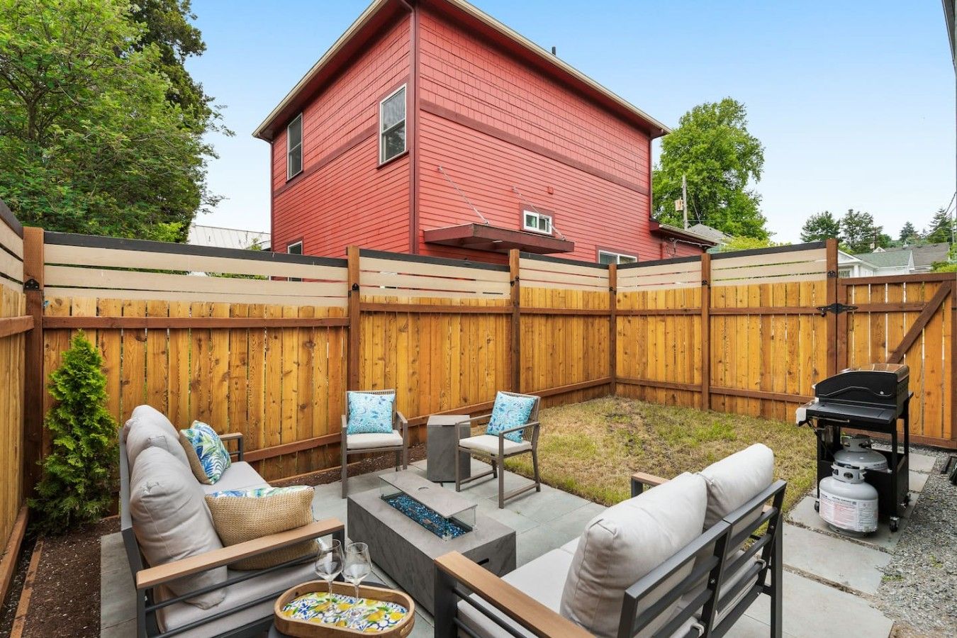 A backyard with a red house, a wooden fence, outdoor furniture, and a barbecue grill.