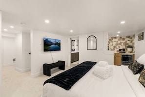Ein modernes Schlafzimmer mit einem großen Bett, einem Fernseher, einer Bank und einem im Hintergrund sichtbaren Badezimmer.