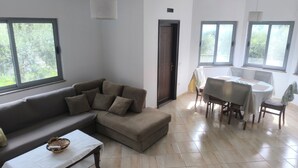 Uma sala de estar com sofá, mesa de jantar e cadeiras, janelas amplas e uma porta.