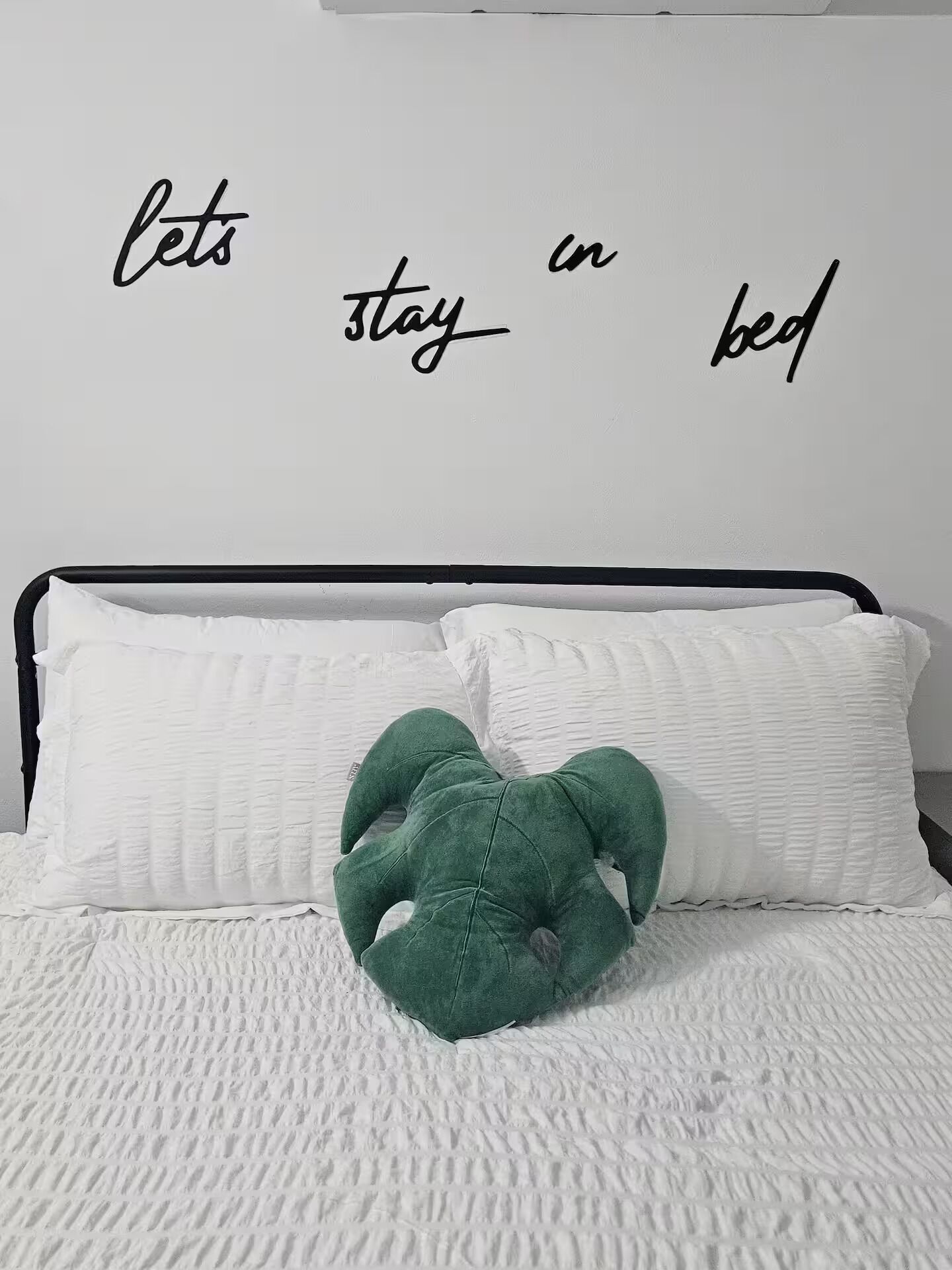 Ein Bett mit weißer Bettwäsche, ein grünes Kissen und eine Wand mit dem Text 'lets stay in bed'.