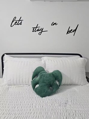Un lit avec une literie blanche, un oreiller vert en peluche et un mur sur lequel on peut lire le texte : « restons au lit ».