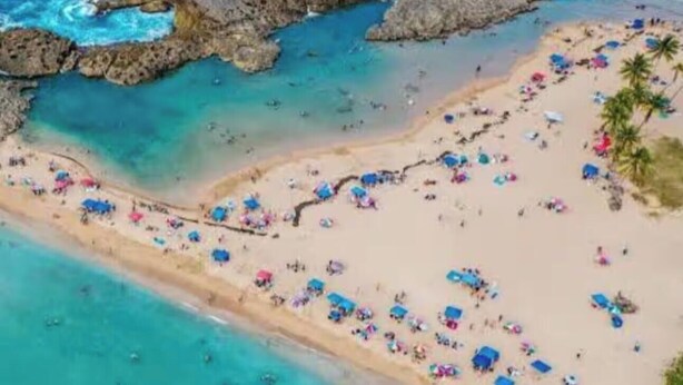 Een overzicht vanuit de lucht van een strand met helder turquoise water, rotsformaties en tal van strandparasols.