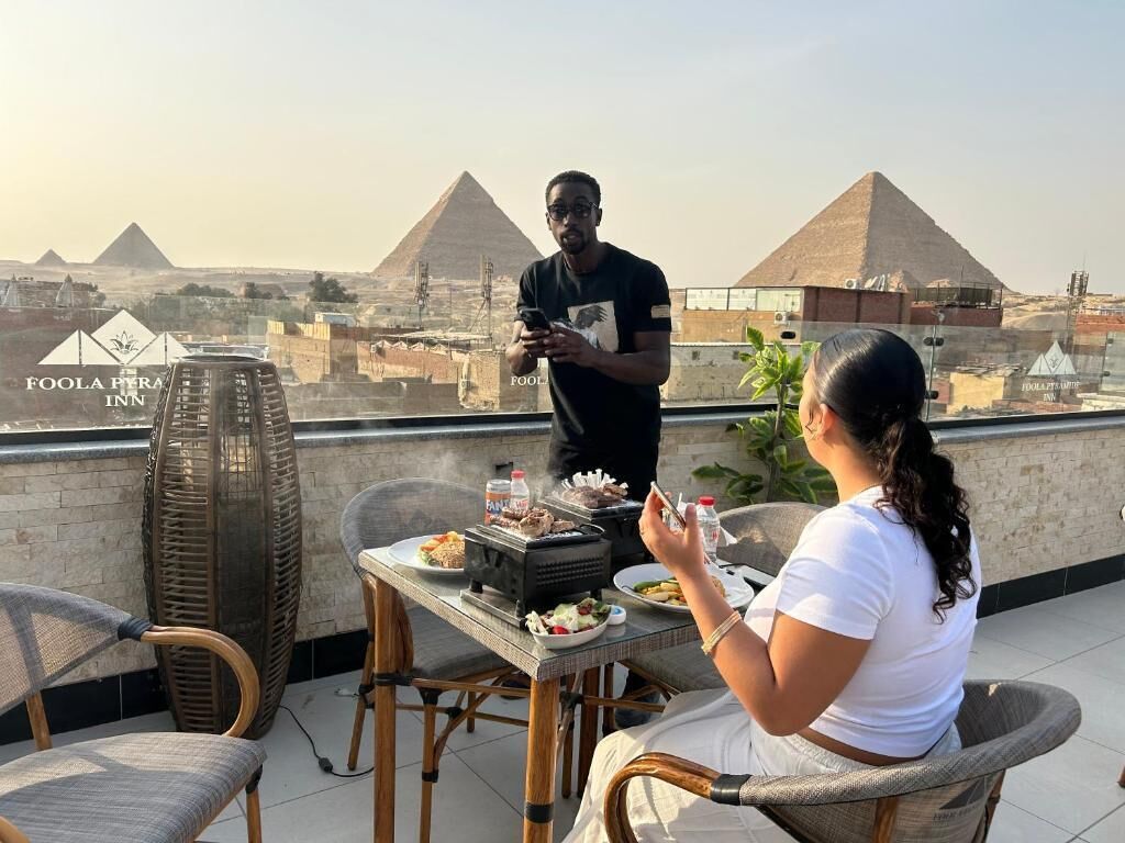 Un espace de restauration aménagé sur le toit, offrant une vue sur les pyramides, une table dressée avec de la nourriture et une personne debout.