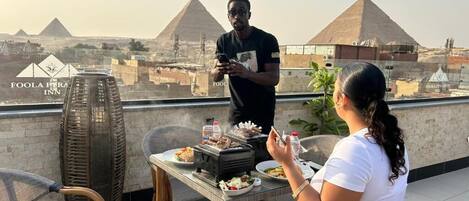 Un espace de restauration aménagé sur le toit, offrant une vue sur les pyramides, une table dressée avec de la nourriture et une personne debout.