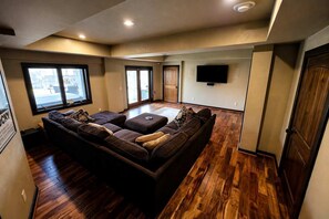 Ein geräumiges Wohnzimmer mit einer großen Eckcouch, Holzboden, einem flachbildfernseher an der Wand und einer Glastür zum Außenbereich.