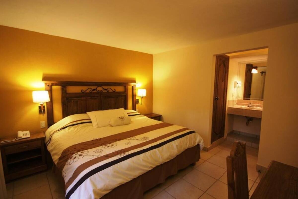 A hotel room with a bed, bedside lamps, a nightstand, a wooden headboard, a bathroom with a sink and mirror, and a wooden chair.