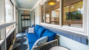 Eine überdachte Veranda mit einem blauen Sofa, einem Korbstuhl und einem Fenster mit Blick ins Freie.