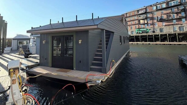 Une péniche est amarrée à un quai, sous un ciel dégagé, entourée d’eau et d’un immeuble doté de nombreux balcons.