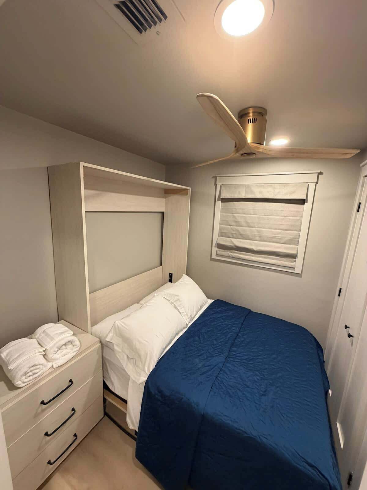 Ein Schlafzimmer mit einem Bett, einem Nachttisch, einer Kommode mit Schubladen, einem Deckenventilator, einem Fenster mit Jalousien und einer Tür.