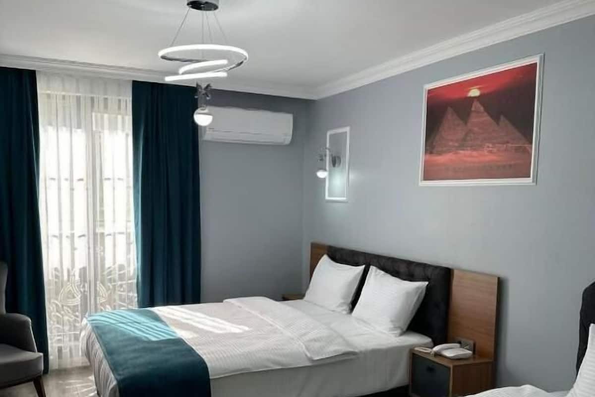 Ein Hotelzimmer mit zwei Betten, einem Sessel, einem Nachttisch, einem an der Wand montierten Telefon und einem Fenster mit Vorhängen.