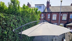 Een terras met een tafel en stoelen, beschut onder een overkapping, omringd door een haag en een bakstenen muur.