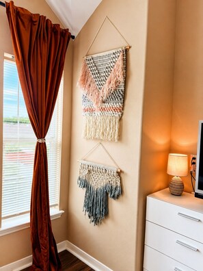 Ein Zimmer mit Fenster, Vorhang, Wandkunst aus Makramee, Lampe und einem weißen Schminktisch.
