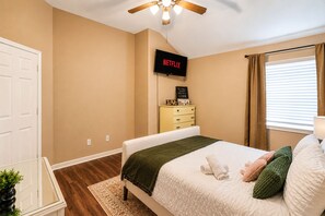 Ein Schlafzimmer mit Bett, Fernseher, Kommode, einem Fenster mit Vorhängen und einem Deckenventilator.