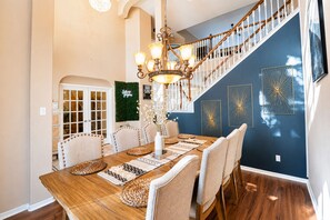 A dining area with a wooden table, beige upholstered chairs, a chandelier, and a staircase with a blue wall and decorative wall art. - FIFA Traveler's Oasis Pool Sauna Patio Arcade Sleep 14+ (Richmond)