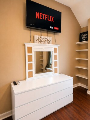 Ein weißer Schminktisch mit Spiegel, ein an der Wand befestigter Fernseher mit dem Schriftzug 'NETFLIX' und ein Schild mit der Aufschrift 'Coffee Break'.