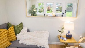 A living room with a sofa, pillows, a white throw blanket, a side table with a lamp, and a mirror reflecting the room. - The Ellery | Garden Suite Historic Queen’s Park (New Westminster)