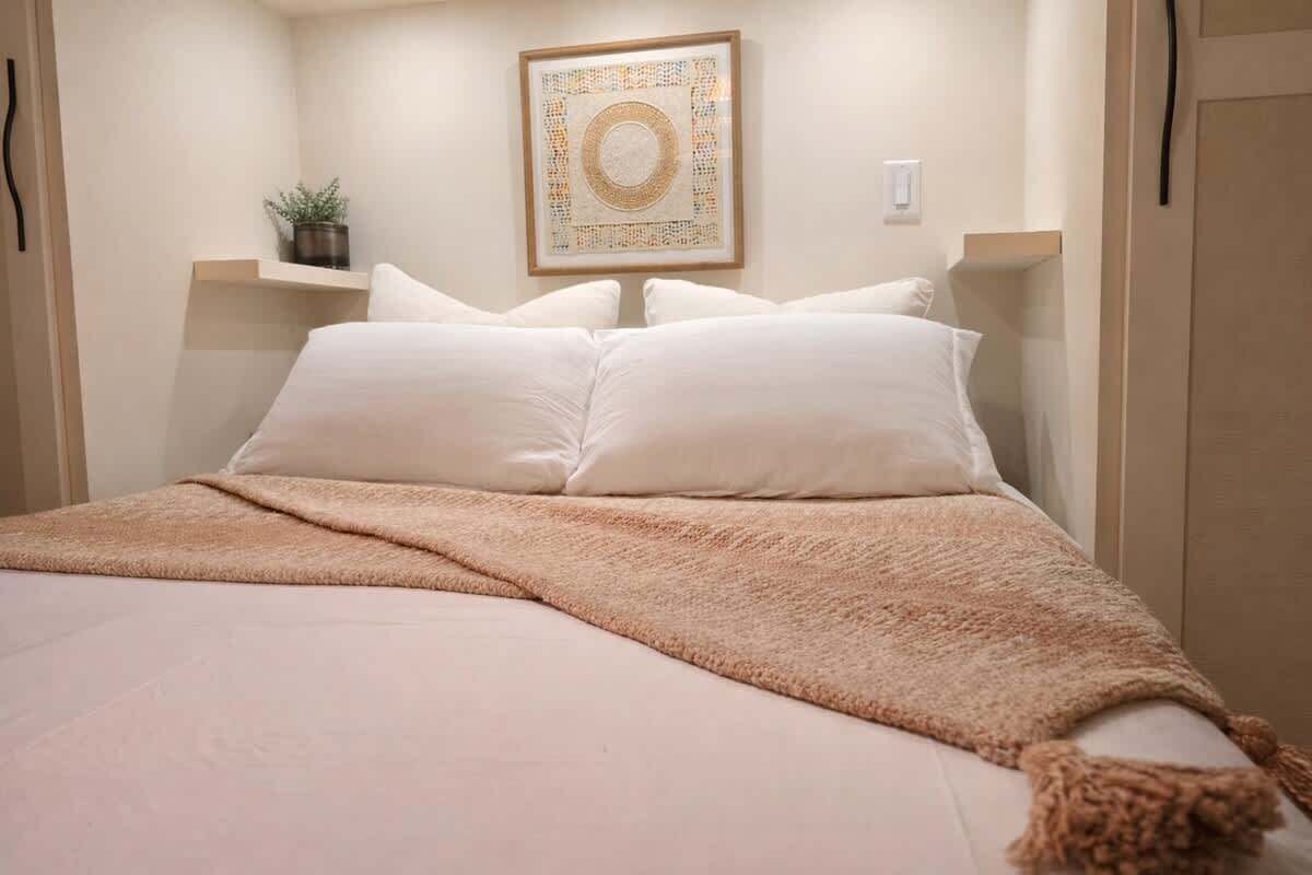 Un lit bien fait, avec du linge de lit blanc et un plaid marron, une œuvre d’art encadrée au mur et une petite plante en pot posée sur une étagère.