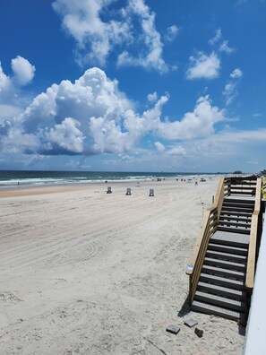 Ein Strand mit einer Holztreppe, die zum Sand hinunterführt, bewölkter Himmel und dem Meer im Hintergrund.