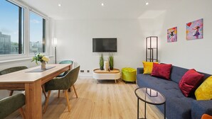 A modern living room with a wooden dining table, green chairs, a blue sofa with colorful cushions, a flat-screen TV, and a large window offering a city view. - Peaceful 1 bed apt in the heart of Fitzrovia (Greater London)
