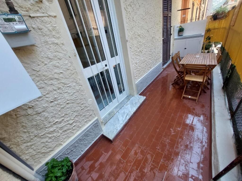 Une terrasse avec un sol carrelé de tuiles rouges, une table et des chaises en bois, une porte blanche avec une fenêtre et une petite plante verte.