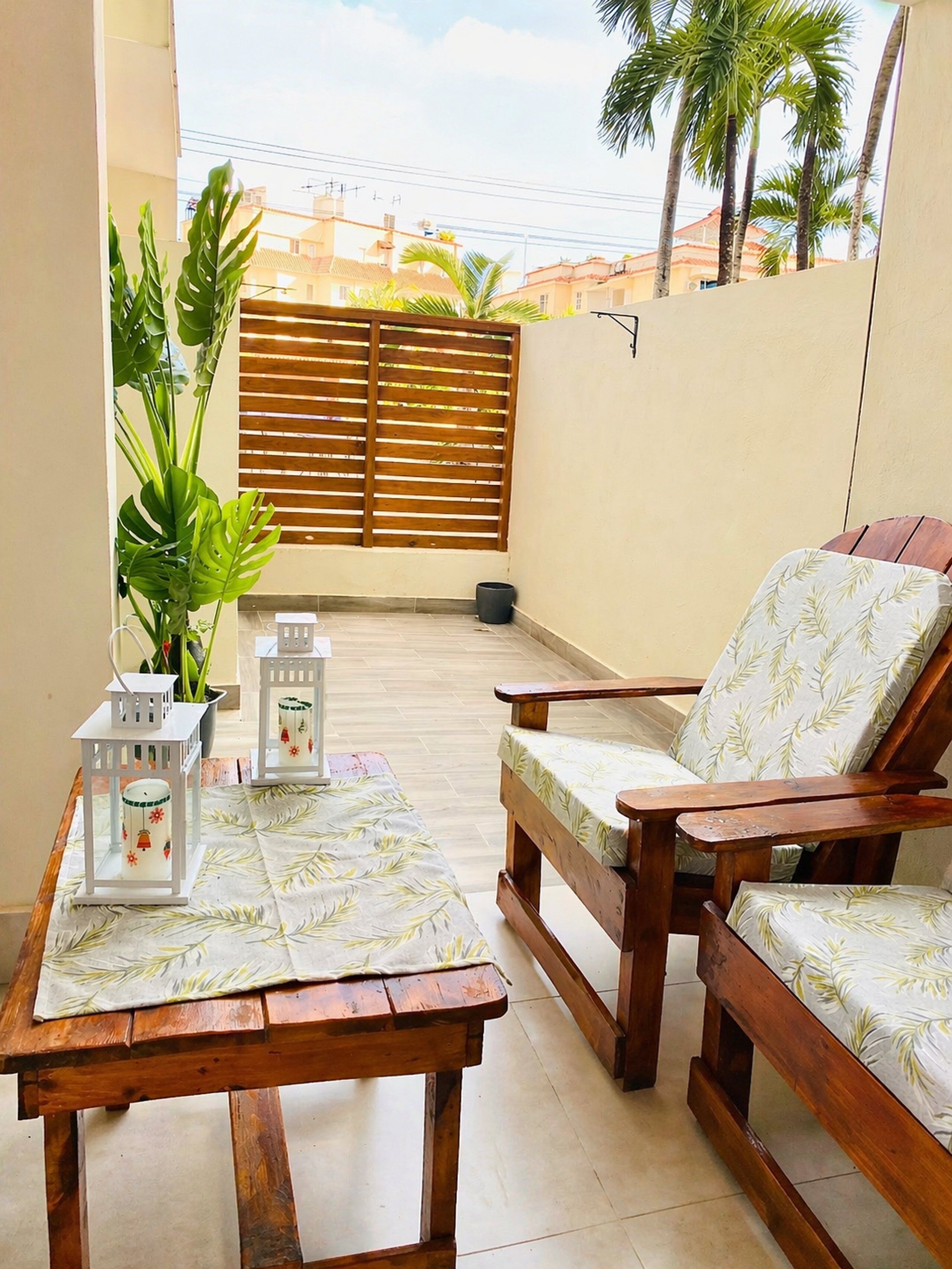 A wooden table with two chairs, a potted plant, and two lanterns on a balcony with a view of palm trees and buildings.