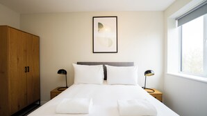 Una cama bien hecha con ropa de cama blanca, un armario de madera, dos mesitas de noche con lámparas y un cuadro en la pared.