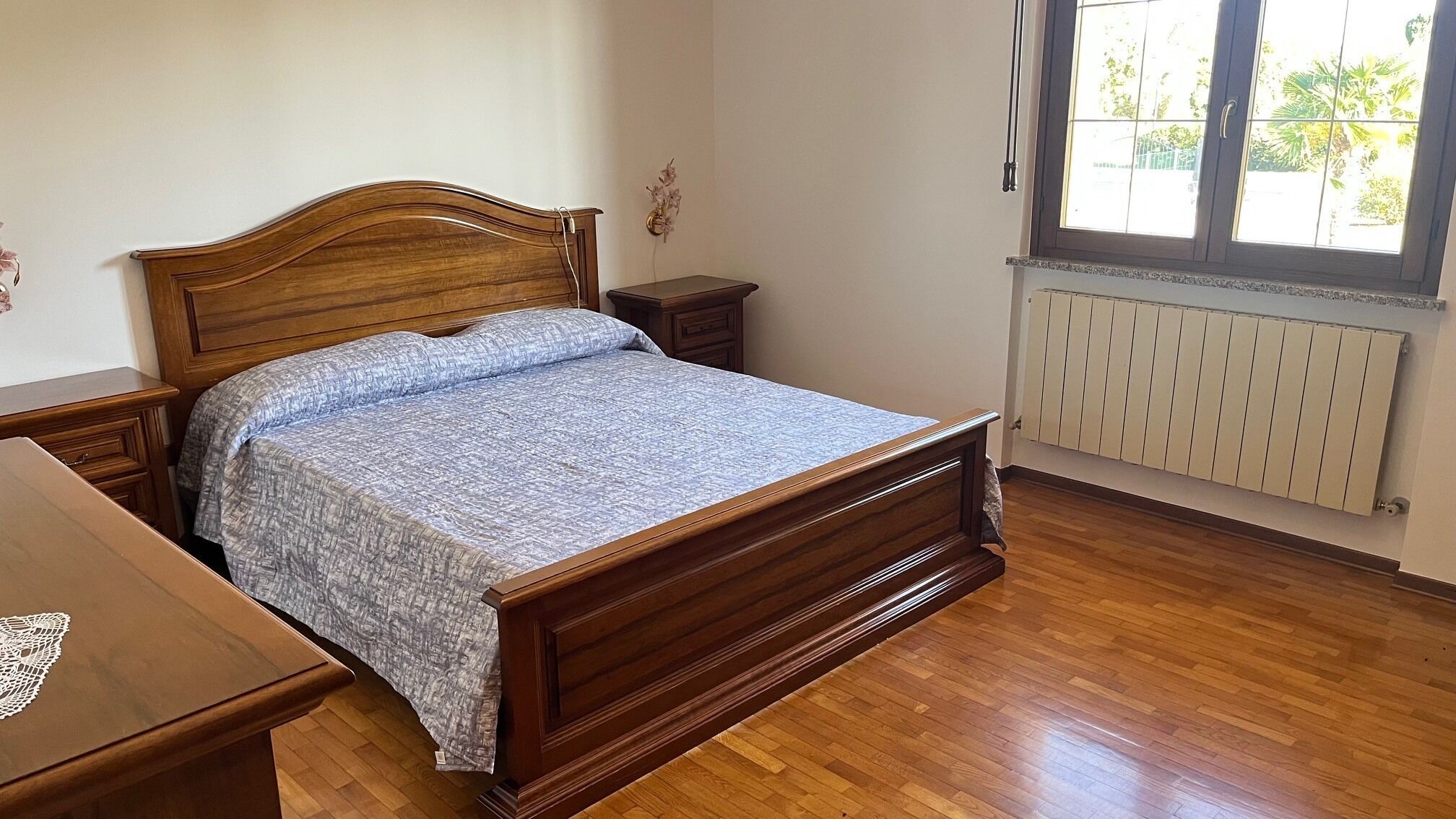 Ein Schlafzimmer mit einem Holzbett, einem Nachttisch, einer Kommode und einem Fenster mit Blick auf Grünflächen.